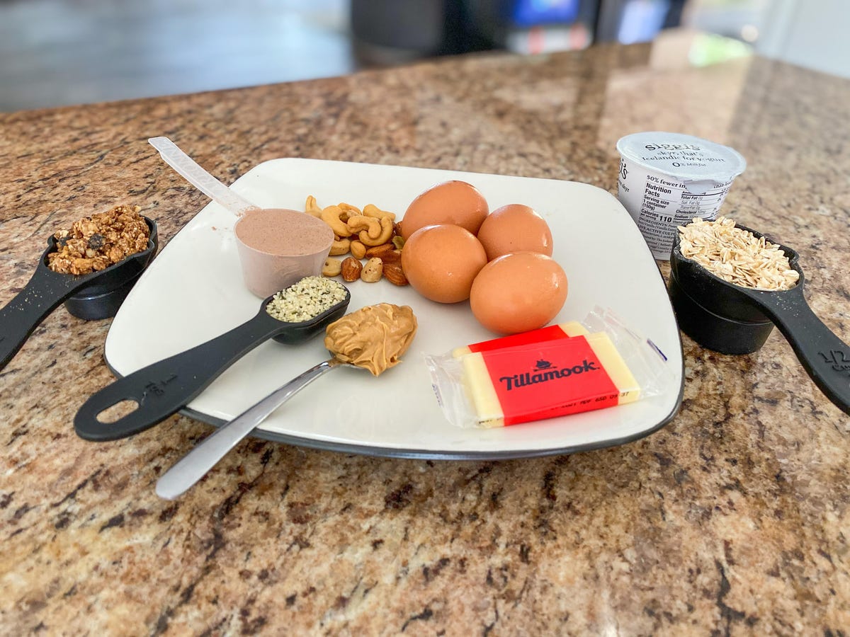 a spread of food containing granola, protein powder, hemp seeds, peanut butter, cheese, nuts, eggs, oats, and yogurt depicting 100 grams of protein