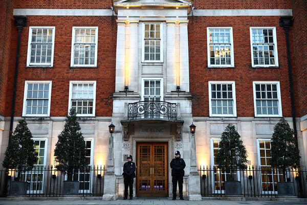Two police officers stand guard beside the entrance to a multistory brick and stone building in London.
