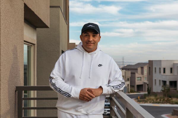 Steve Hawthorne, wearing a white Nike track suit and a dark baseball cap, is on a balcony in a residential area. 